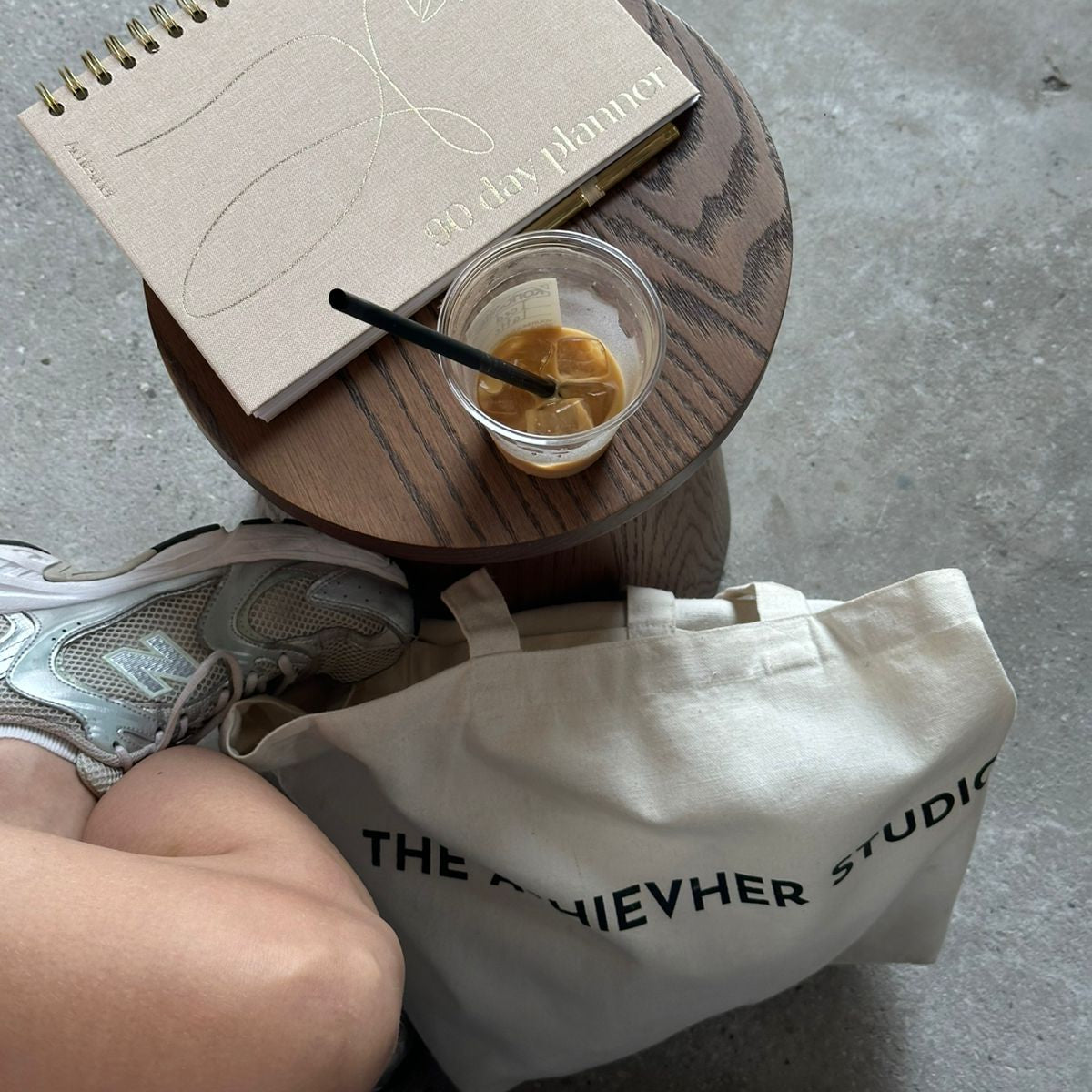 Small round wooden table with a notebook, cup, and bag on a concrete floor.